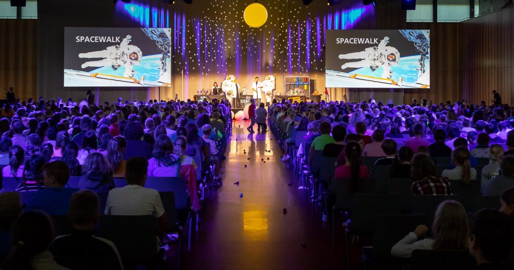 Blick durch das Publikum auf eine Bühne mit Spacewalk-Leinwänden