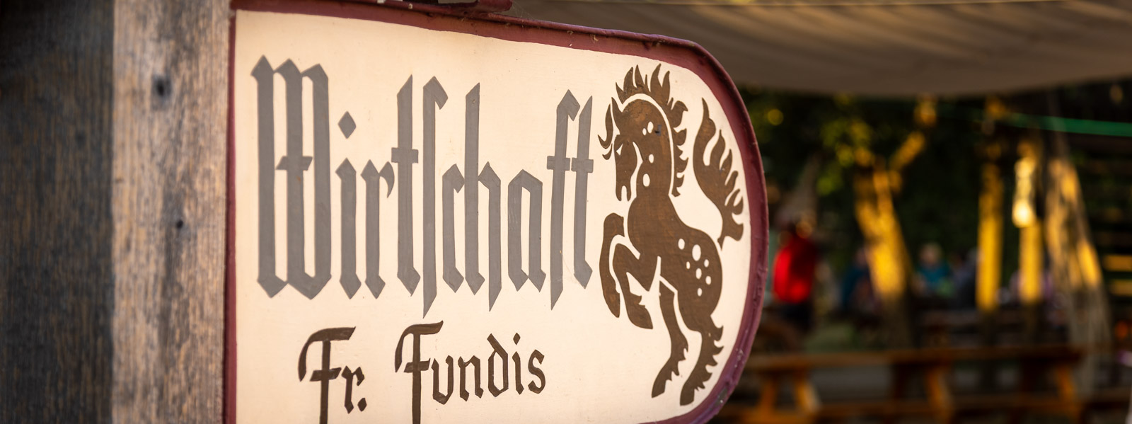 Handgemaltes Wirtschaftsschild „Fr. Fundis“ mit springendem Pferd, dahinter der Biergarten im Abendlicht