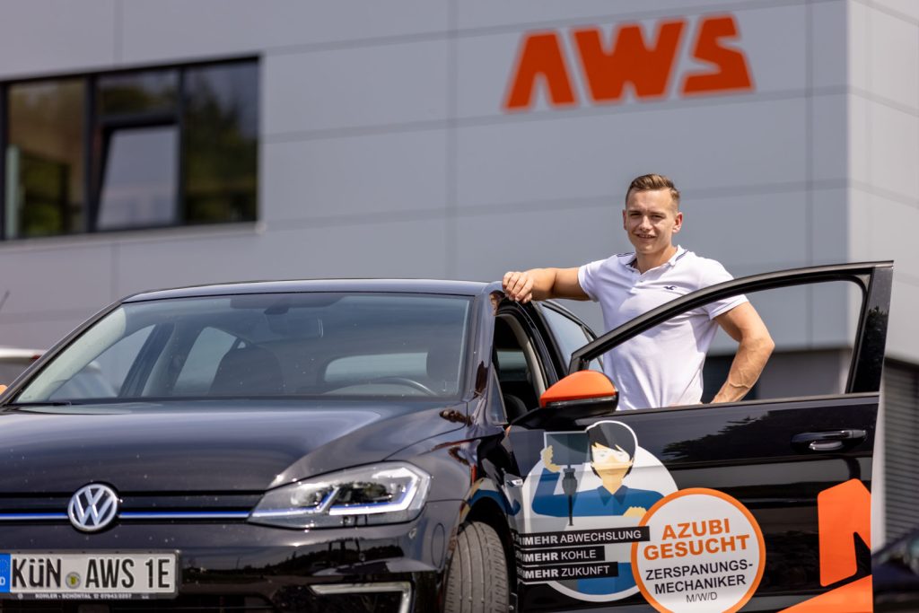 Mitarbeiter lehnt an einem Firmenwagen mit Azubi-Werbung vor einem Firmengebäude