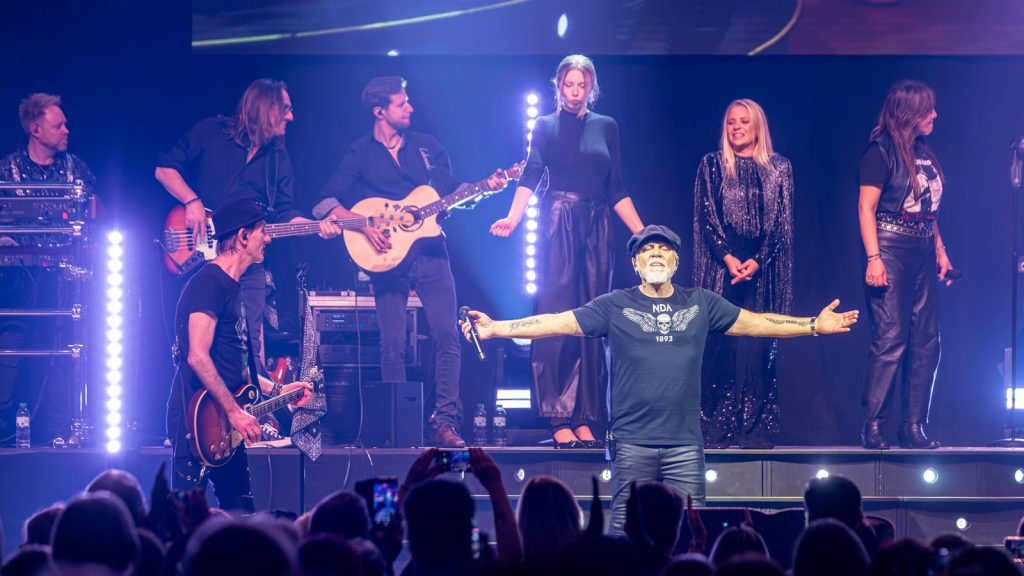 Sänger mit Band auf einer blau beleuchteten Bühne, Arme ausgebreitet vor dem Publikum